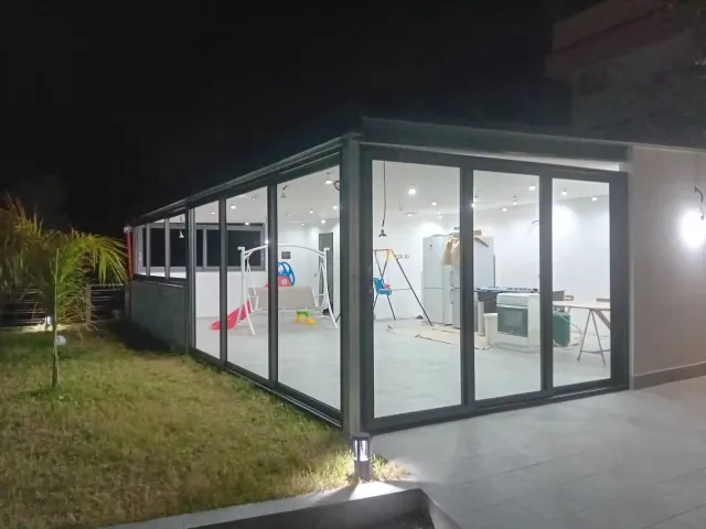 Veranda con pareti in vetro di notte, che illumina l'interno con luce bianca. L'esterno è caratterizzato da prato e patio.