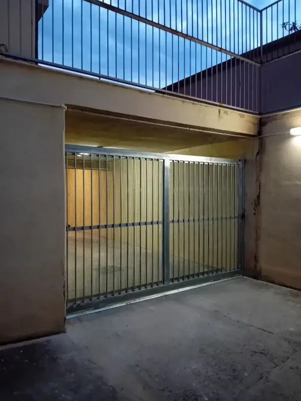 Cancelli metallici sotto una sporgenza di cemento con un balcone sopra, illuminati da una luce calda, esterno al crepuscolo.