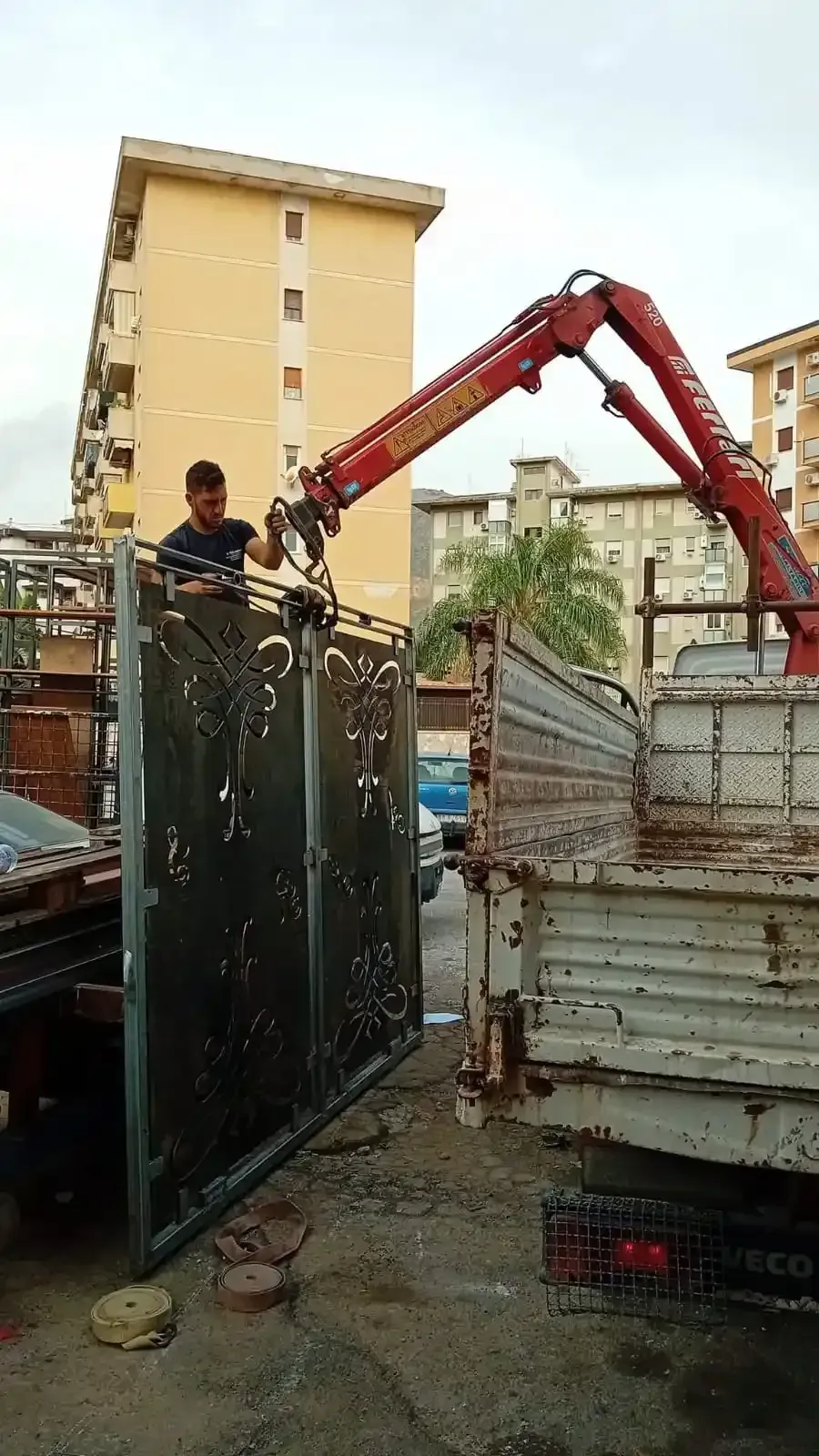 Un operaio utilizza una gru montata su un camion per sollevare un grande cancello metallico sul cassone di un camion.