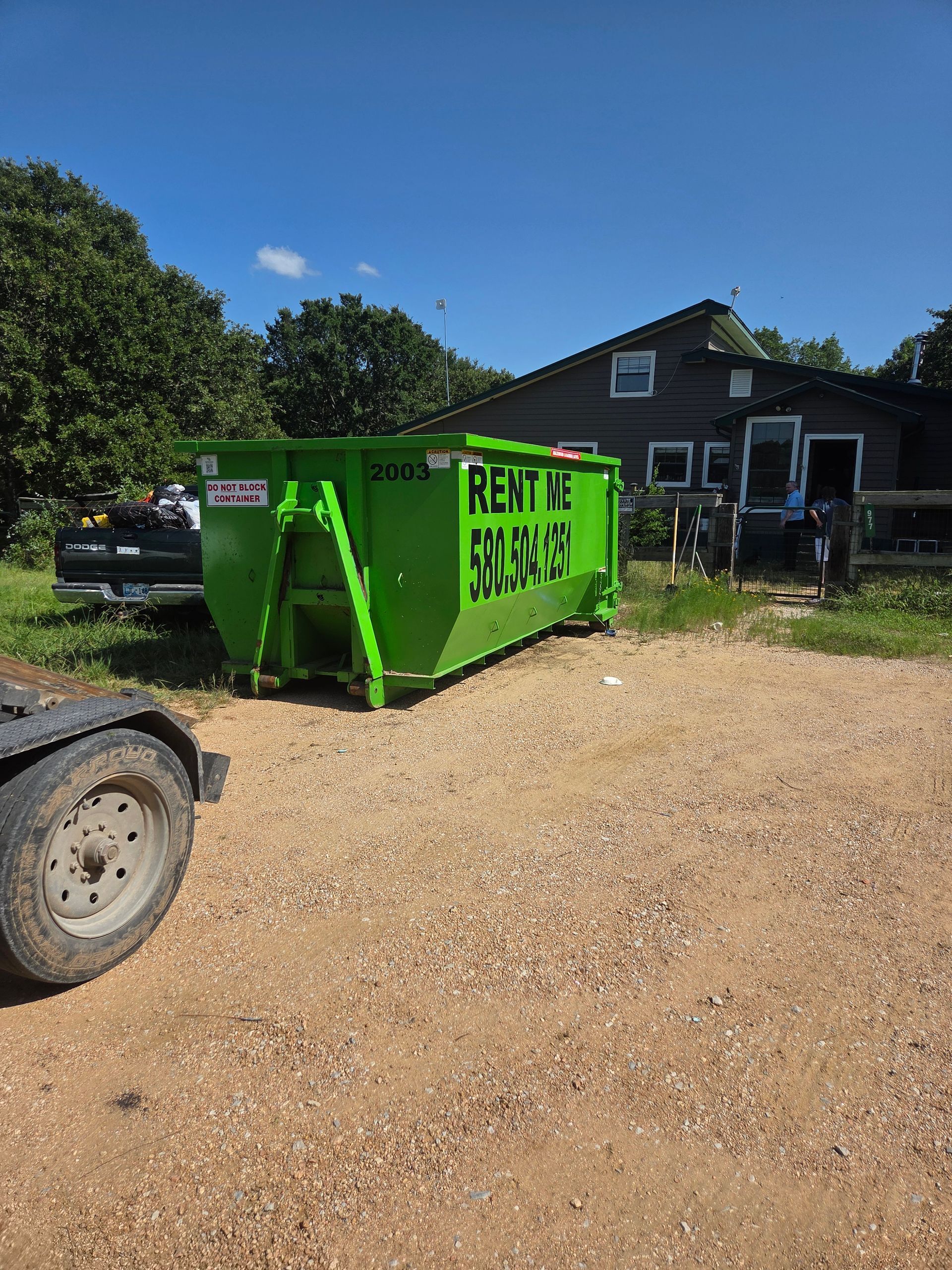 Green rental dumpster on a driveway in front of a brown house, black truck and trees behind.