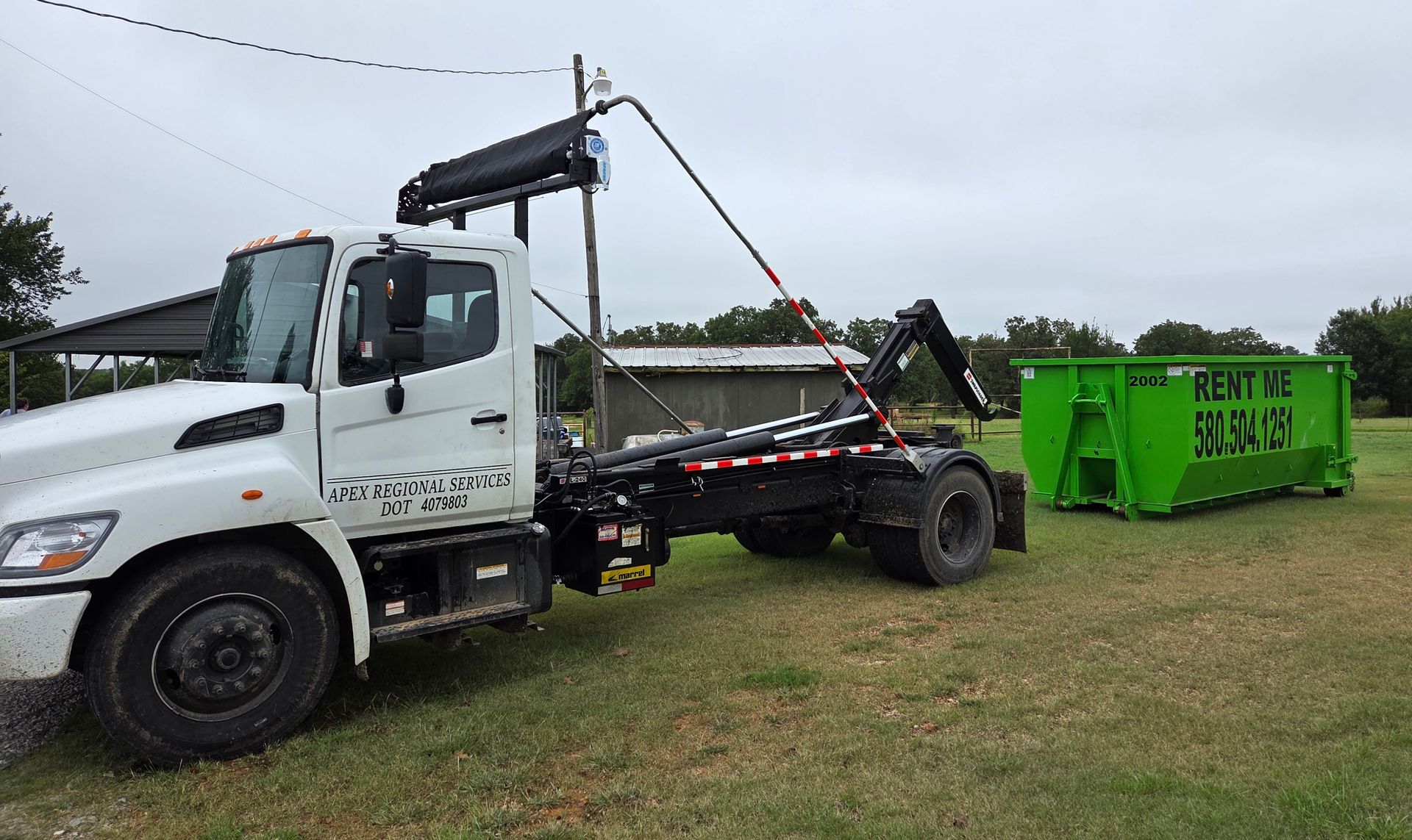 White truck loading a green dumpster on grass in a rural area.