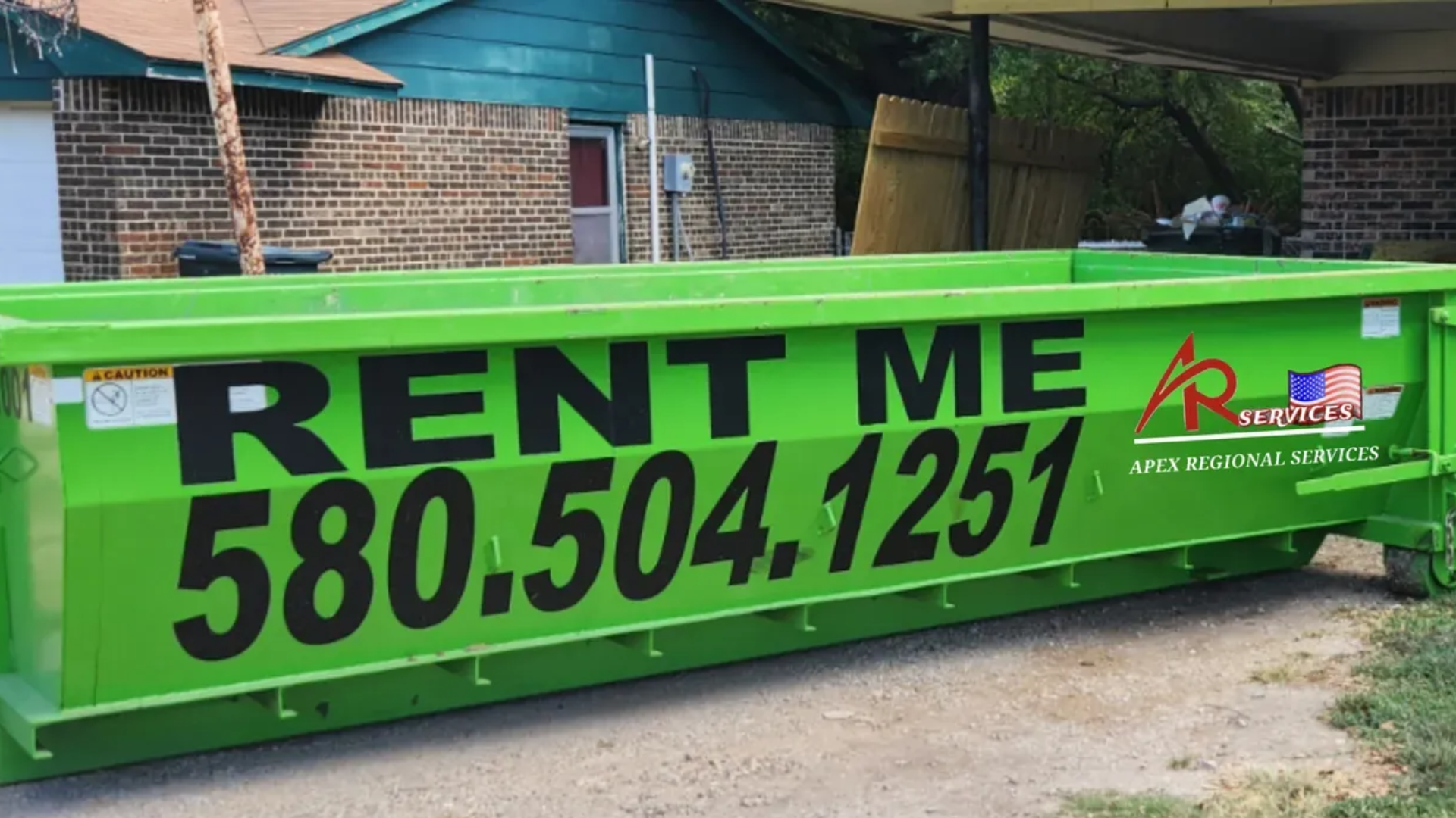 Two green Apex Regional dumpster rentals on a trailer in a grassy area with a building and blue sky in the background.