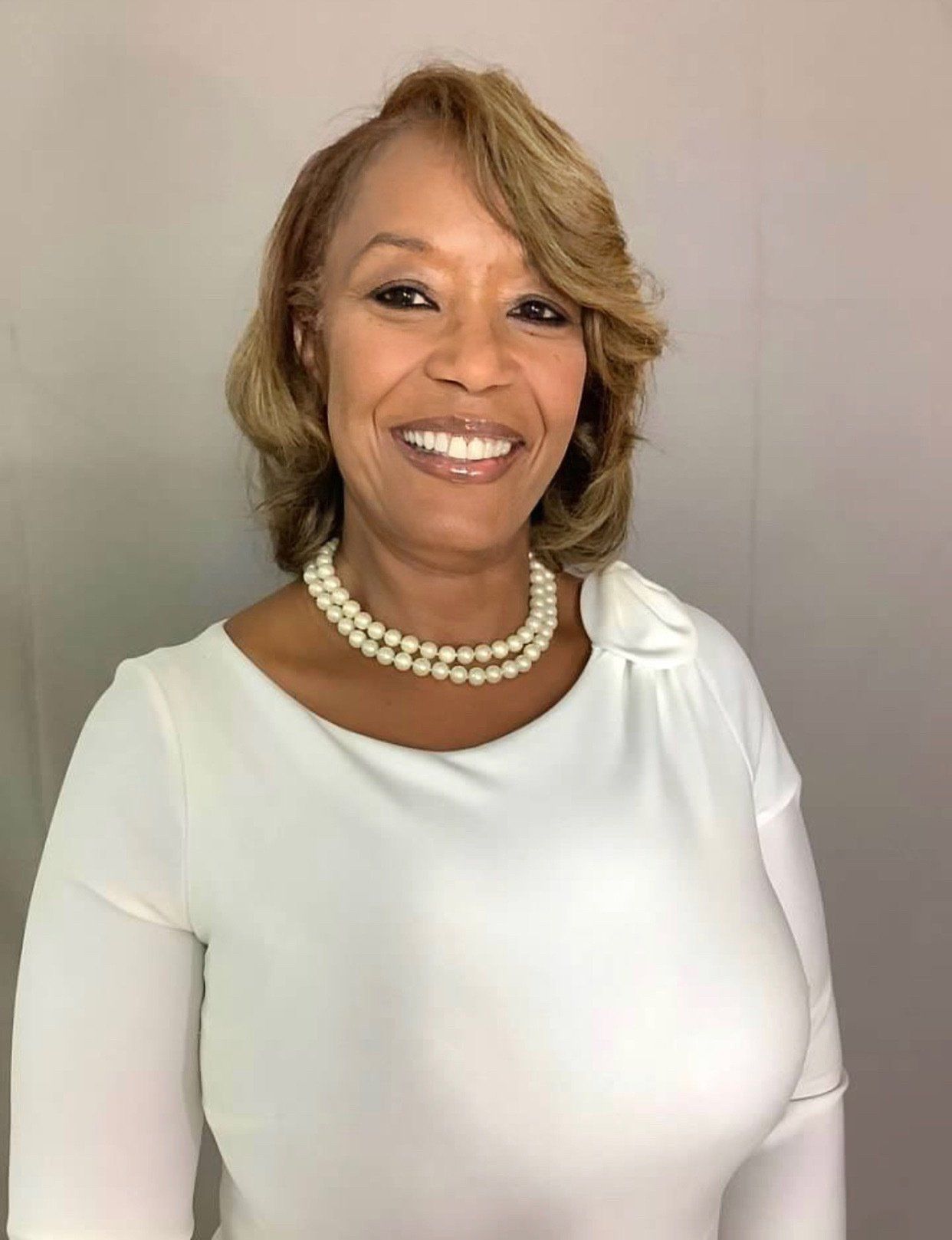 A woman wearing a white dress and a pearl necklace is smiling for the camera.