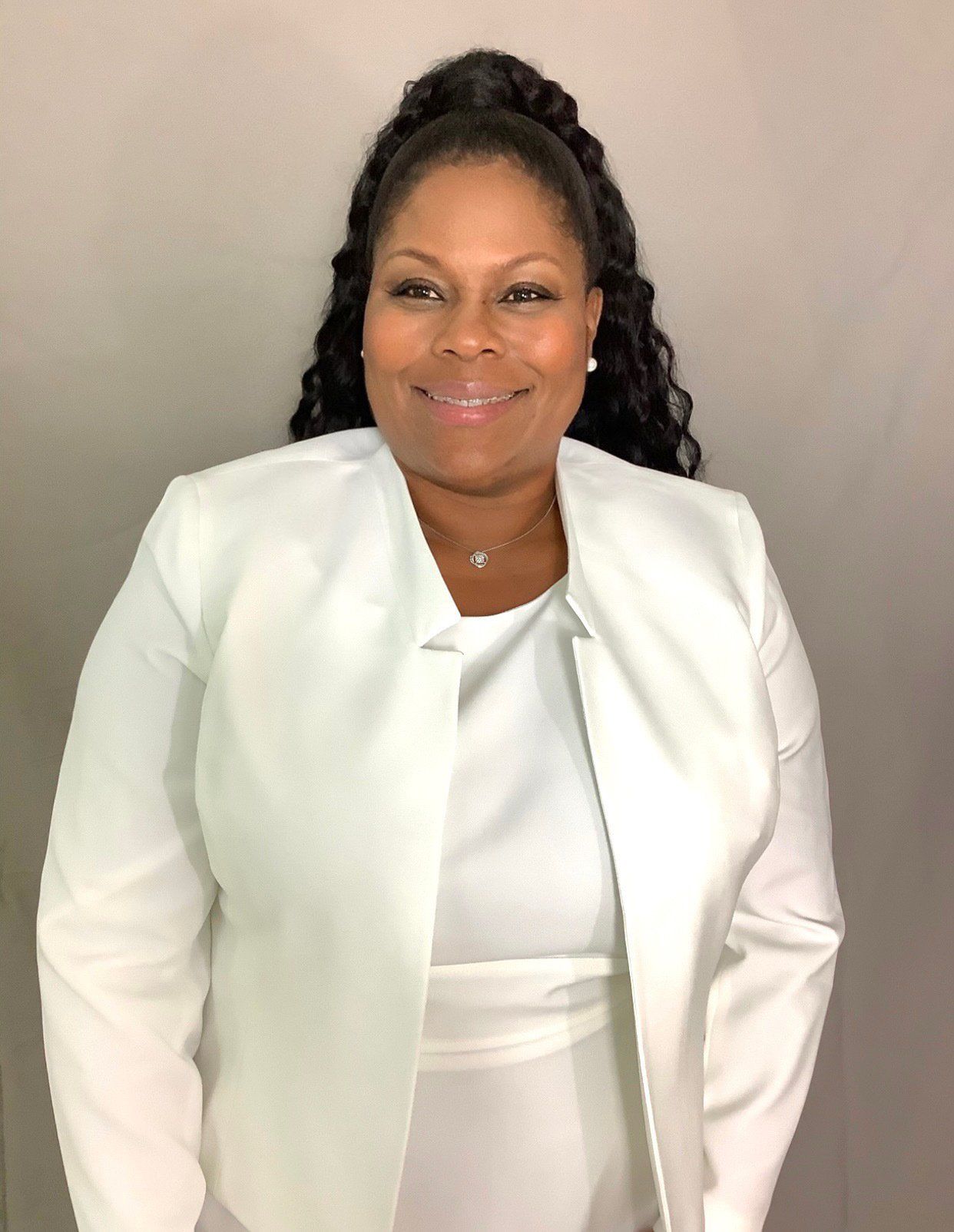 A woman wearing a white jacket and a white dress is smiling for the camera.