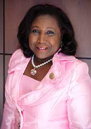 A woman in a pink jacket and pearl necklace is smiling for the camera.