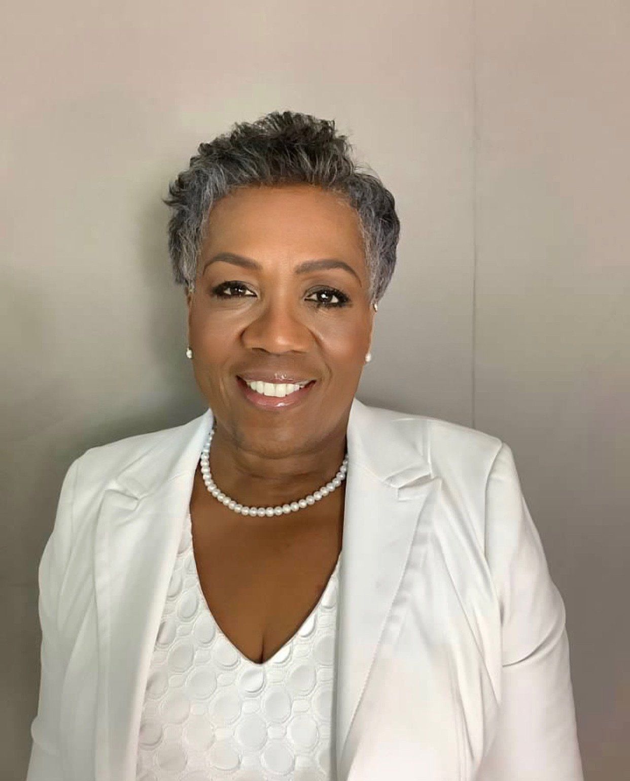 A woman wearing a white jacket and pearl necklace is smiling for the camera.