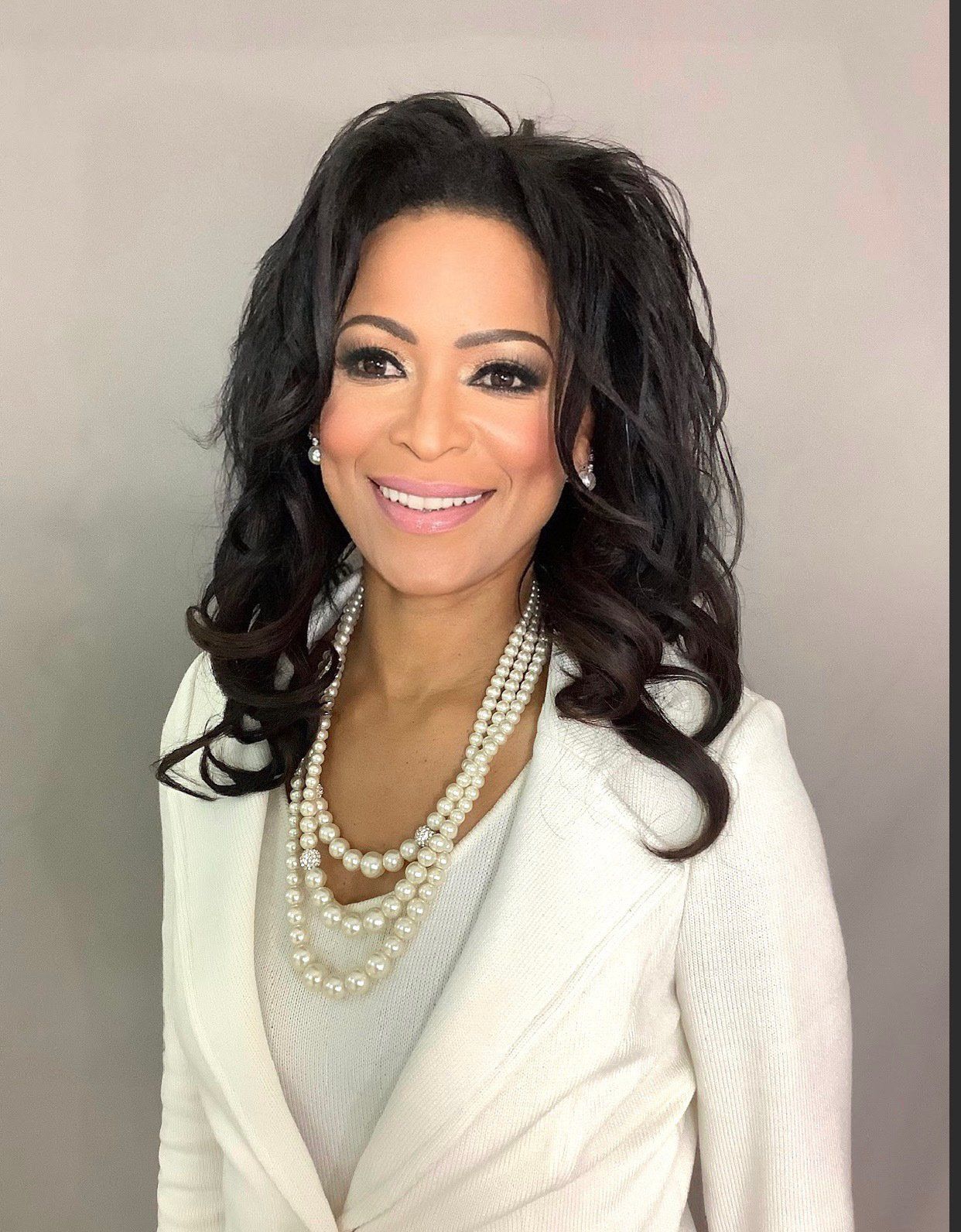 A woman wearing a white jacket and pearls is smiling for the camera.