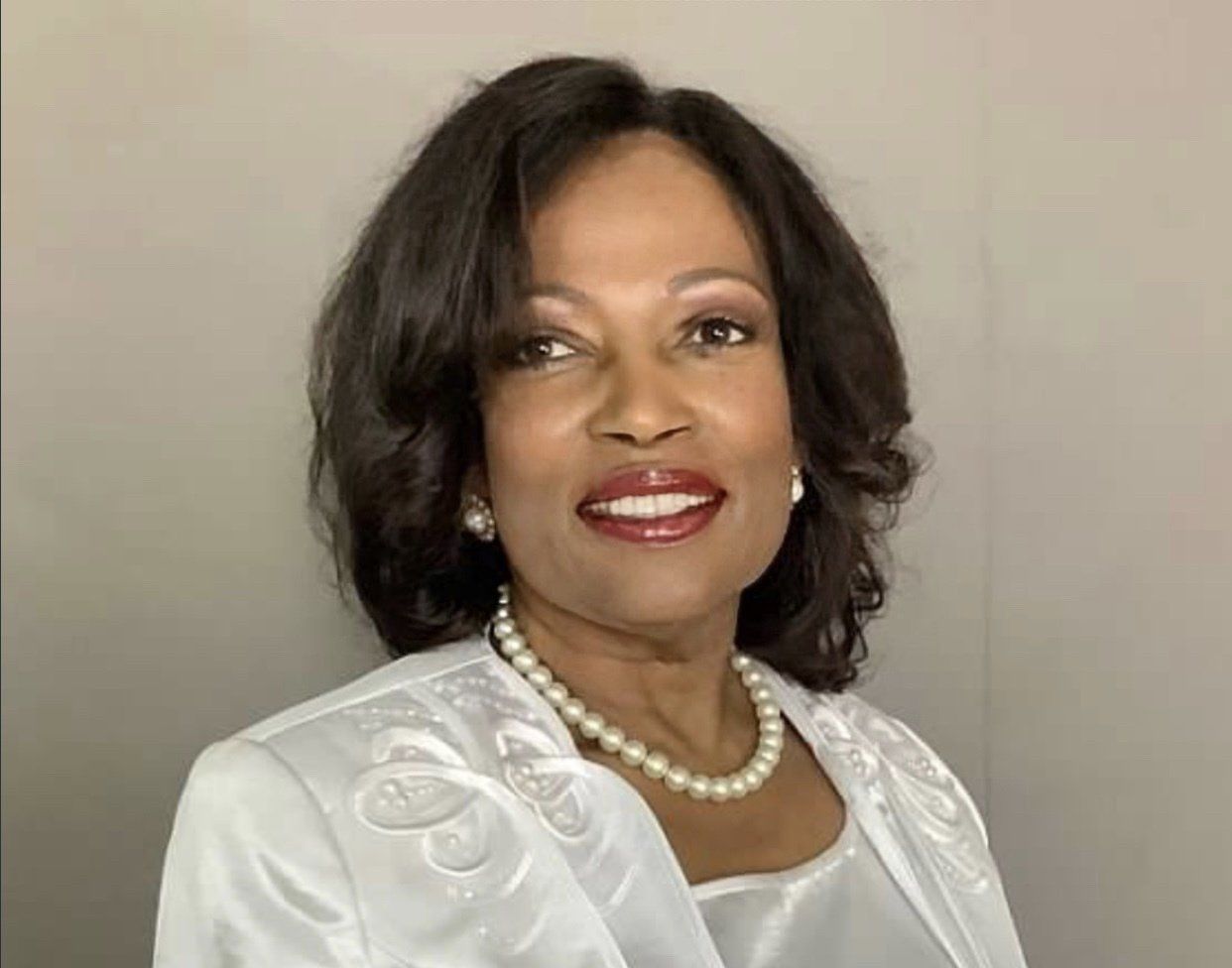 A woman wearing a white jacket and a pearl necklace is smiling.