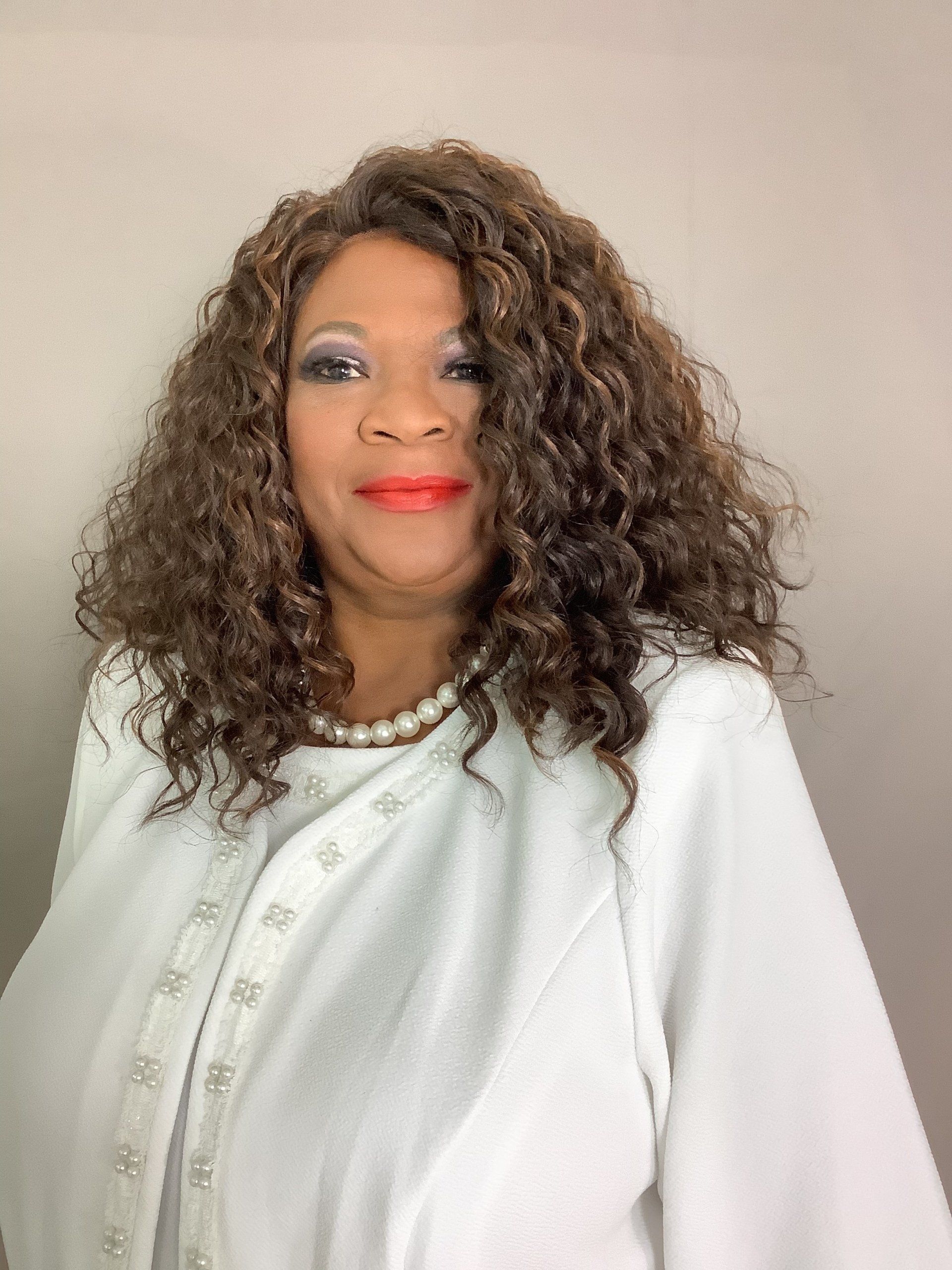 A woman with curly hair is wearing a white jacket and pearls.