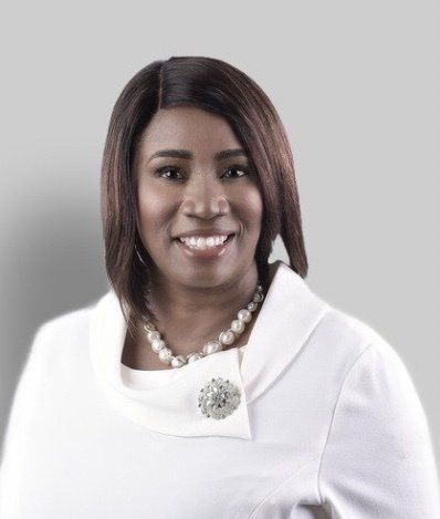 A woman wearing a white jacket and a pearl necklace is smiling.