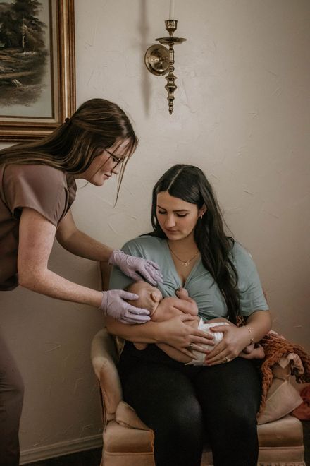 Woman breastfeeding a baby while another woman examines the baby in a light-filled room.