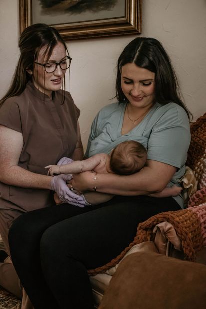 Woman breastfeeding a baby, assisted by a person in brown scrubs. Both women smiling, indoors.