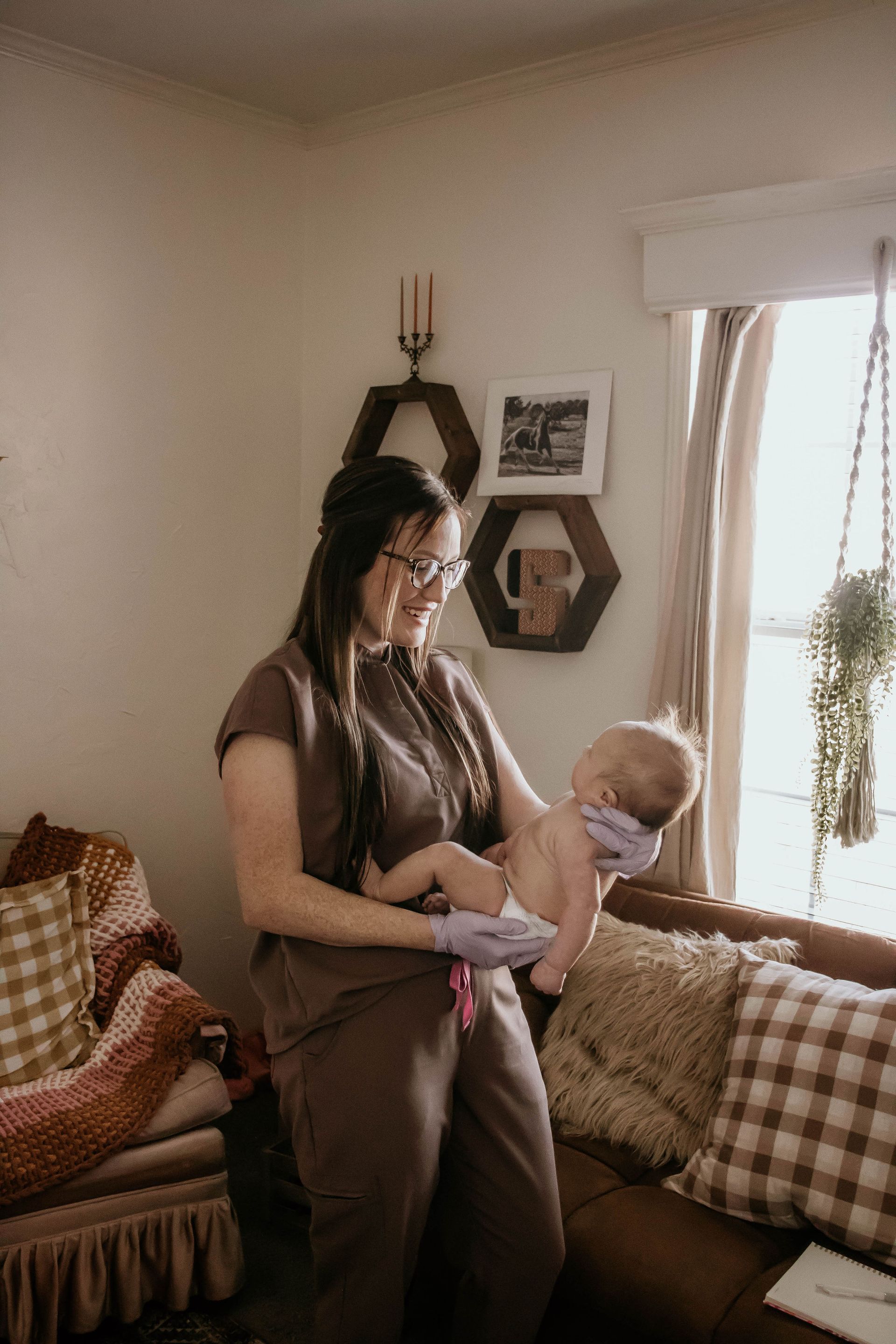 Woman in scrubs holds a newborn baby in a living room, smiling.