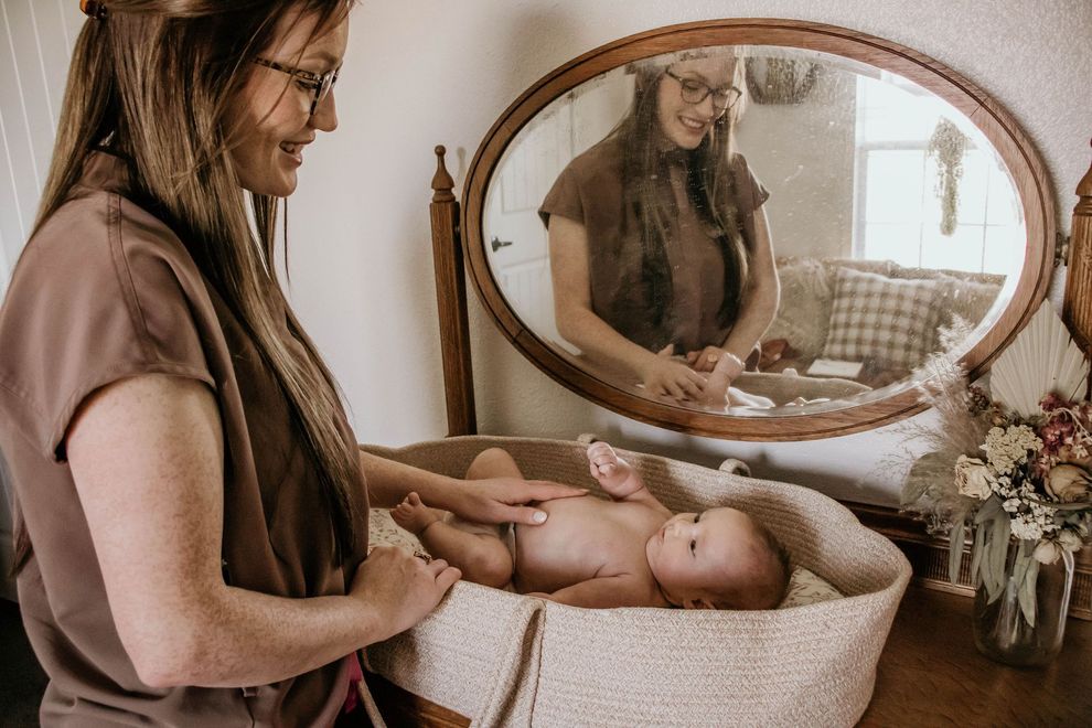 Woman smiles, touching baby in a woven bassinet near a mirror. Warm-toned room, flowers on table.