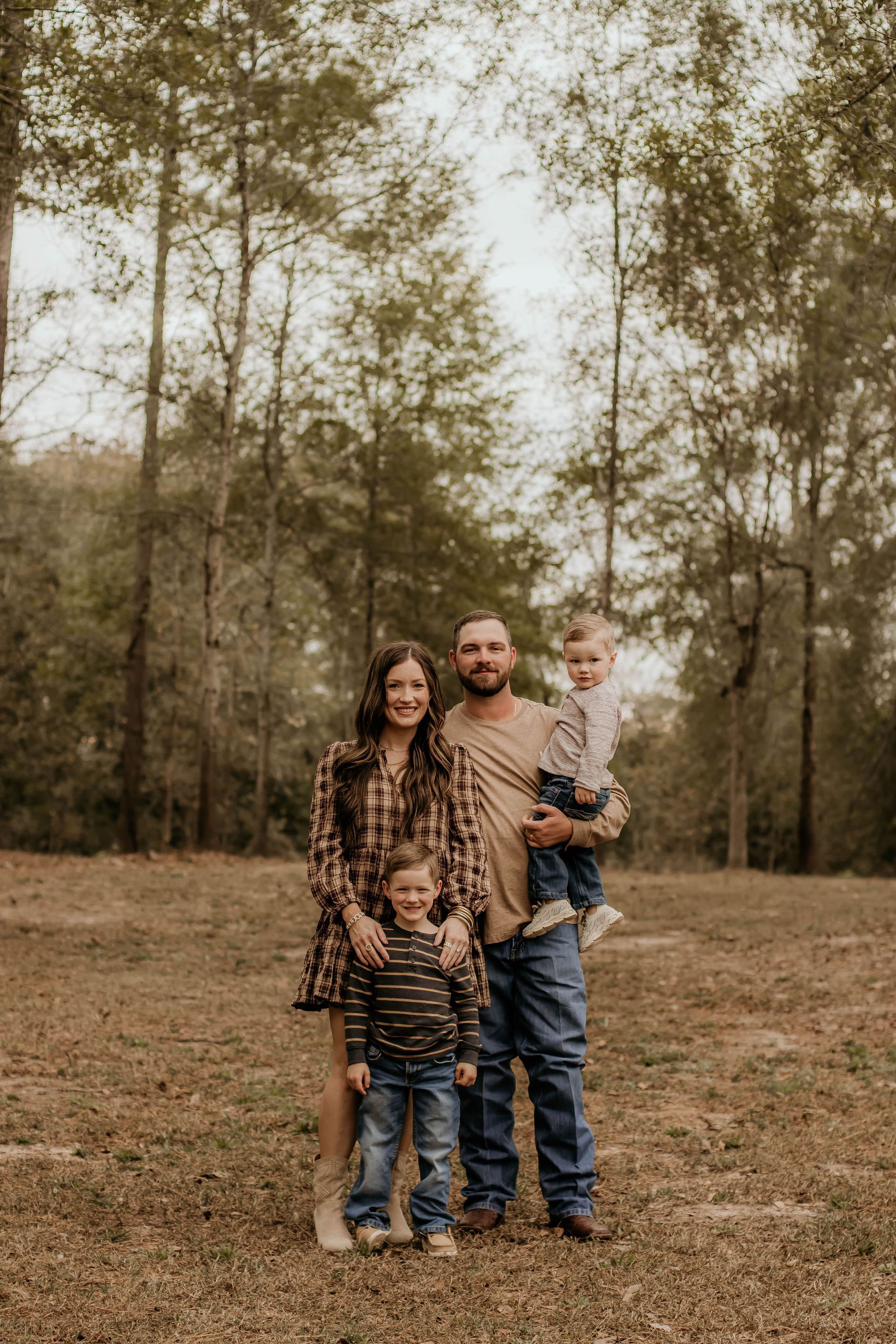 Family of four poses outdoors in front of trees. Woman in plaid, man holding a child, and another child stands in front.