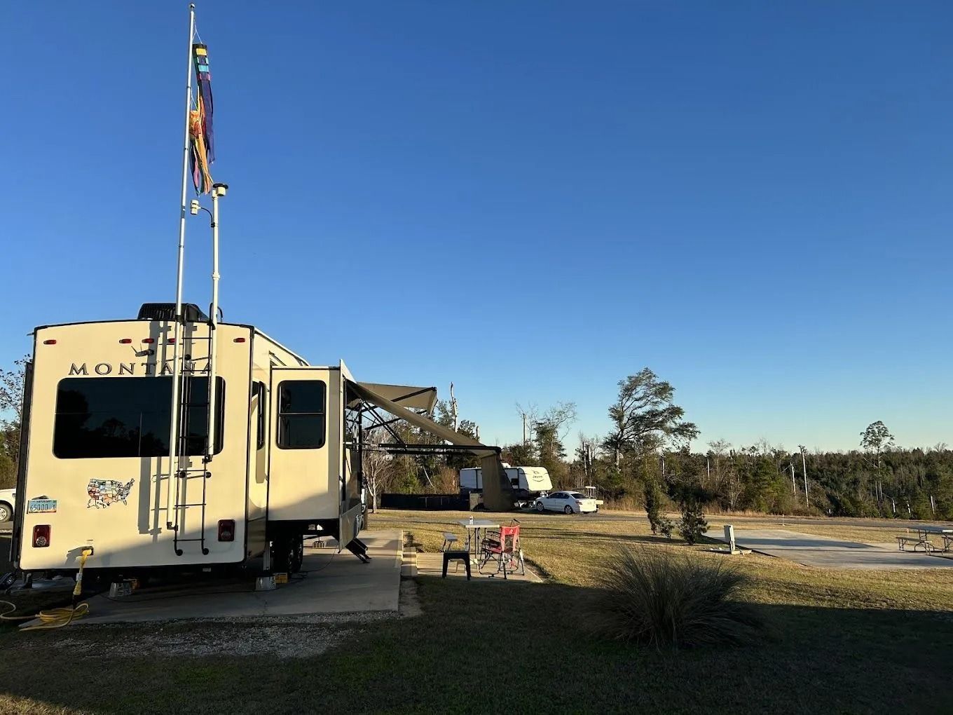 RV campsite with a trailer parked on a concrete pad, flags flying. Clear, blue sky.