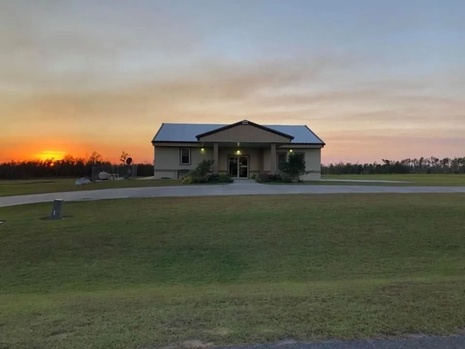 A single-story building with a metal roof sits on a green lawn at sunset; orange and yellow sky.