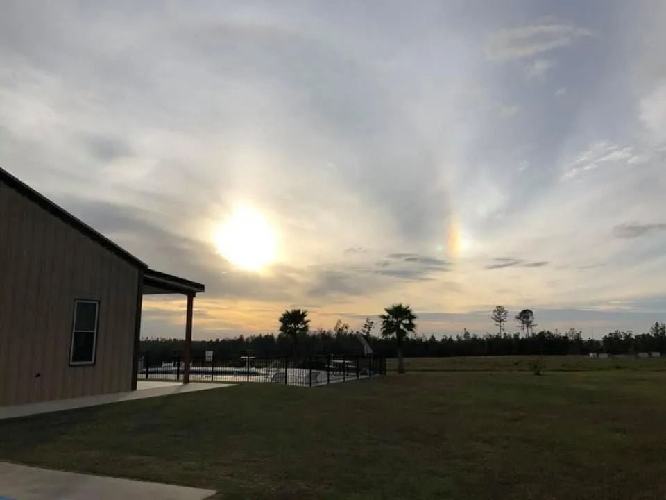 Sun with a sundog display in sky over a building and grassy field.