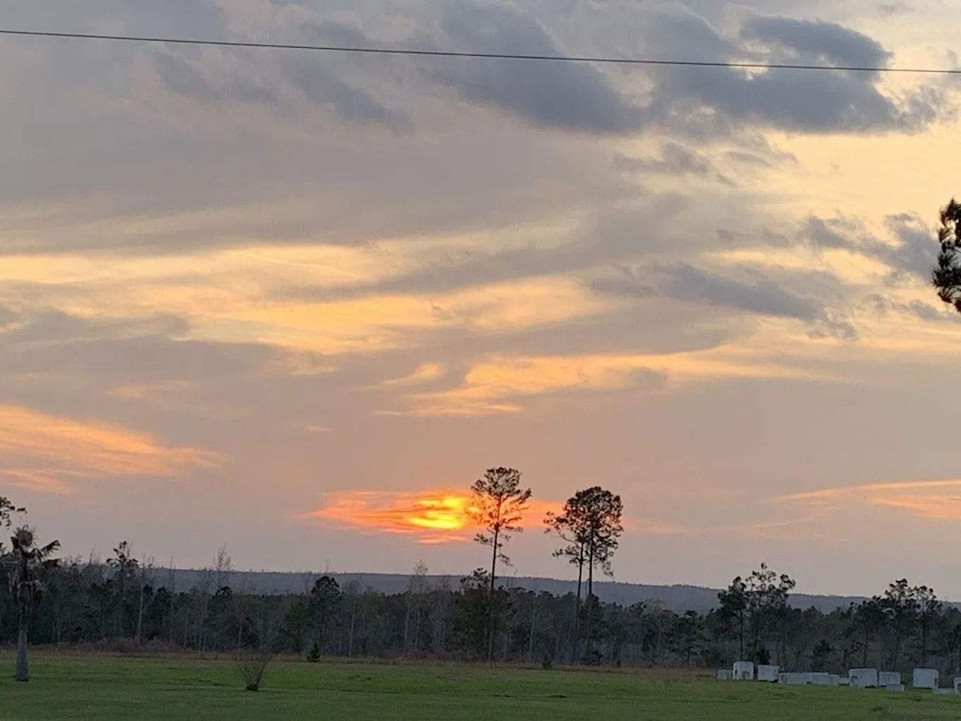 Sunset over a field, with silhouetted trees against a cloudy sky colored in shades of orange and gray.