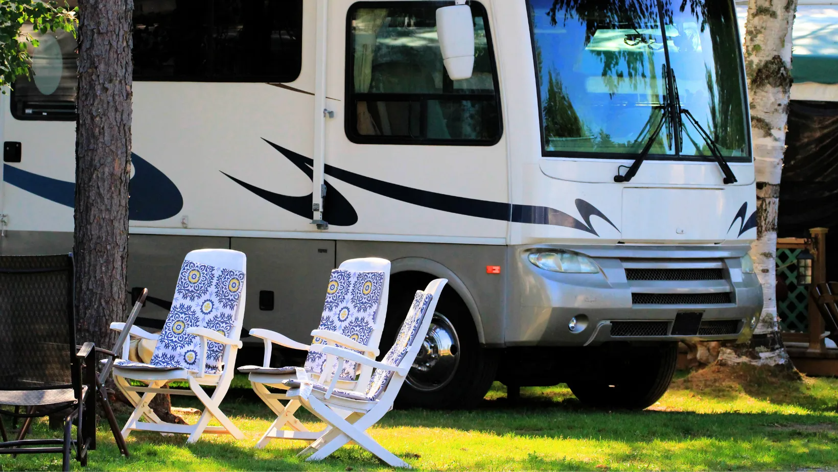 RV parked in grassy area with two white folding chairs.