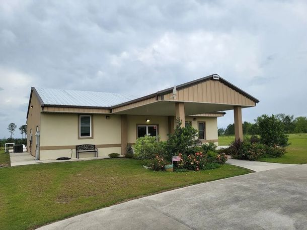 Tan building with metal roof and porch, surrounded by greenery, on a cloudy day.