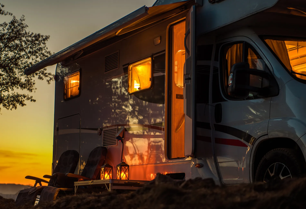 RV at dusk, lit interior, awning open, chairs nearby, golden sunset.