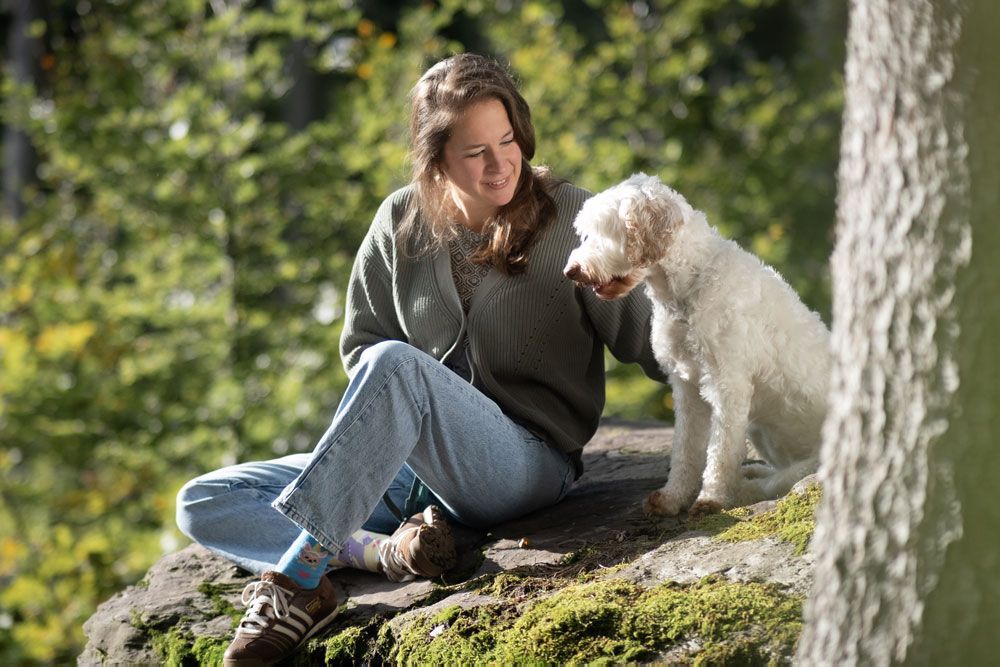 Eine Frau sitzt draussen auf einem Felsen und schaut lächelnd zu einem weissen Hund, den sie sanft berührt.