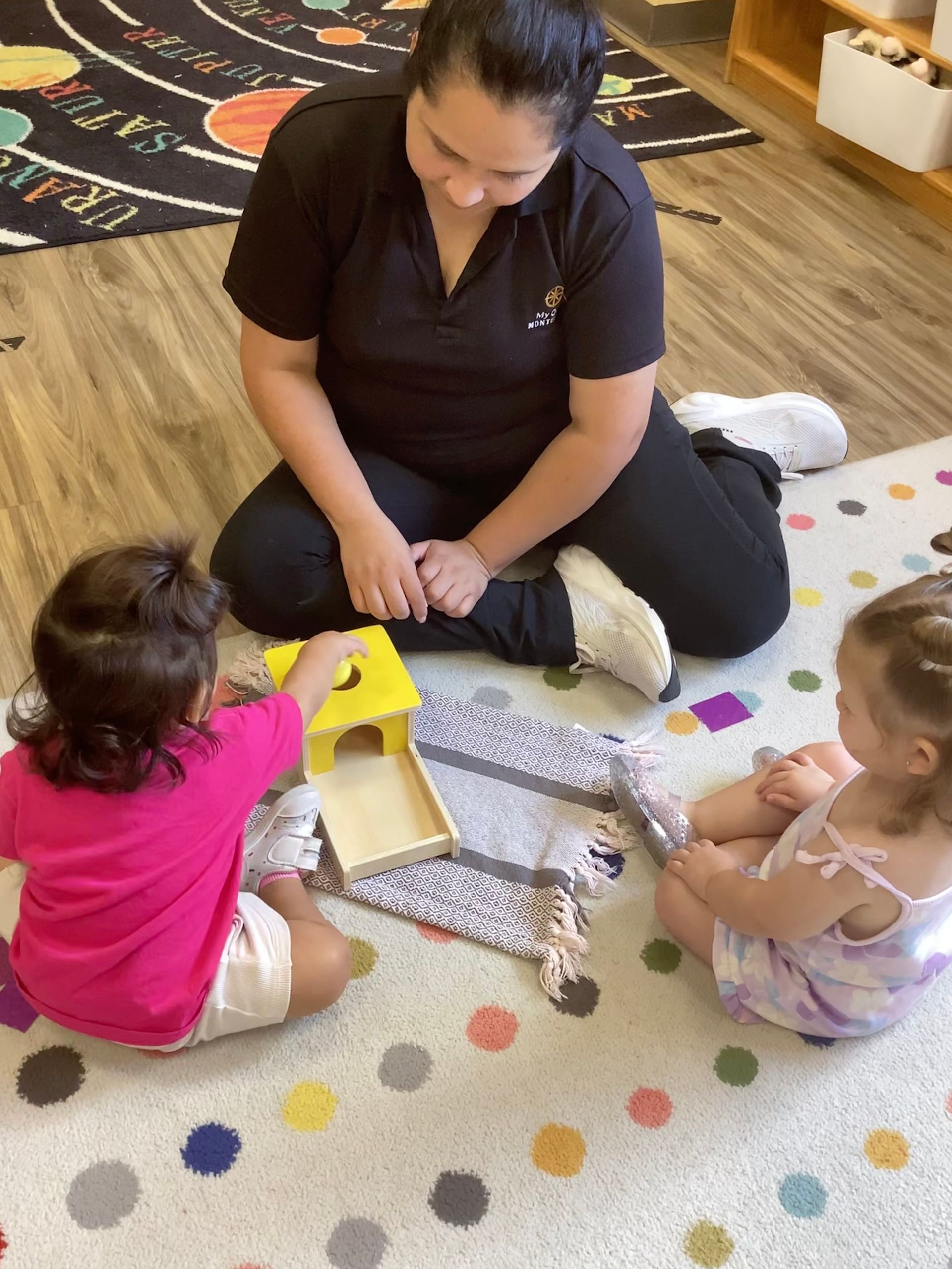 Adult and two toddlers sitting on a play mat, playing with a small toy house and blocks.