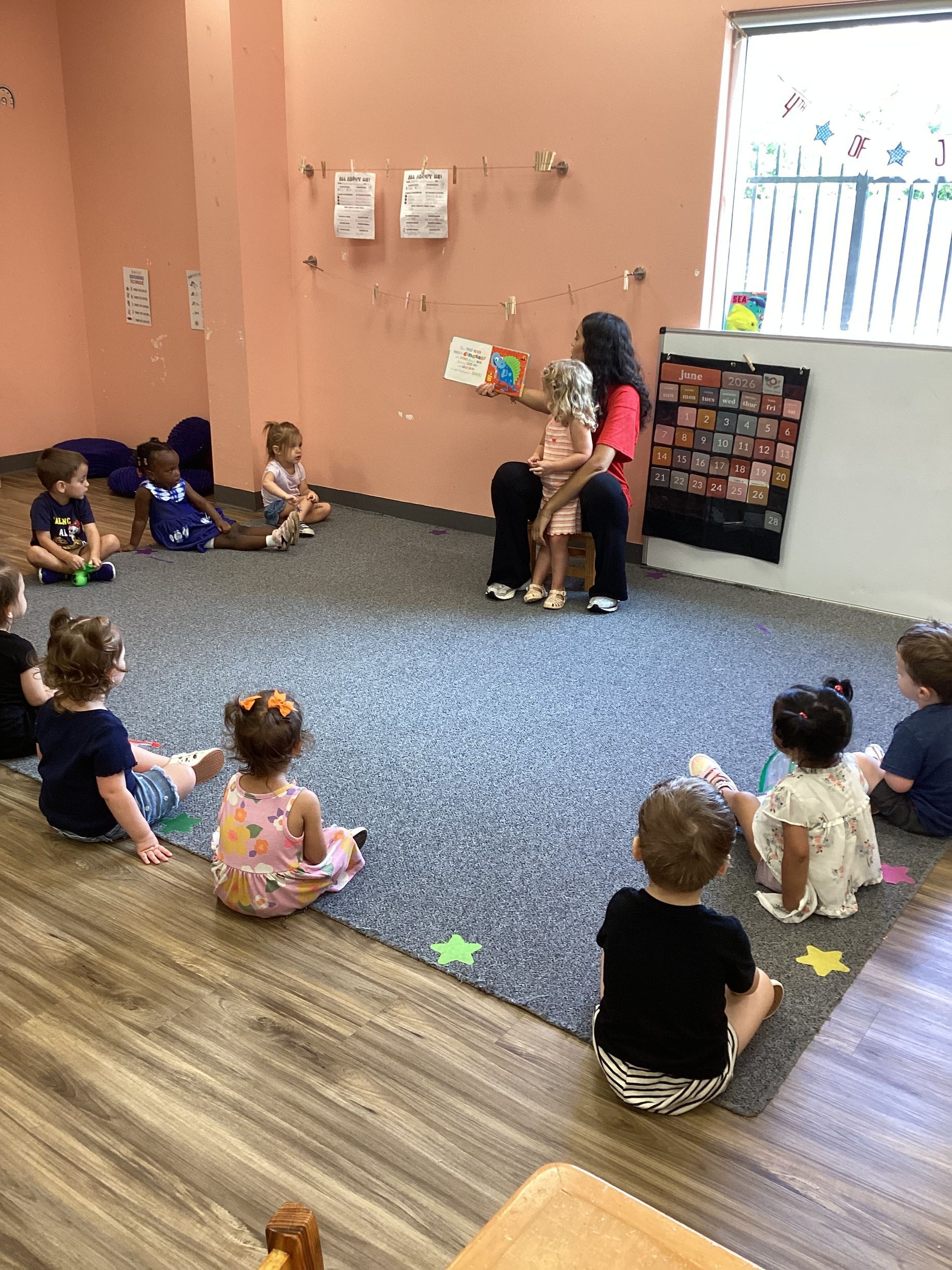 Children seated on the floor in a classroom, watching an adult near a wall display.