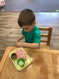 Child seated at a wooden table, looking down at a lunch tray with food in a classroom setting