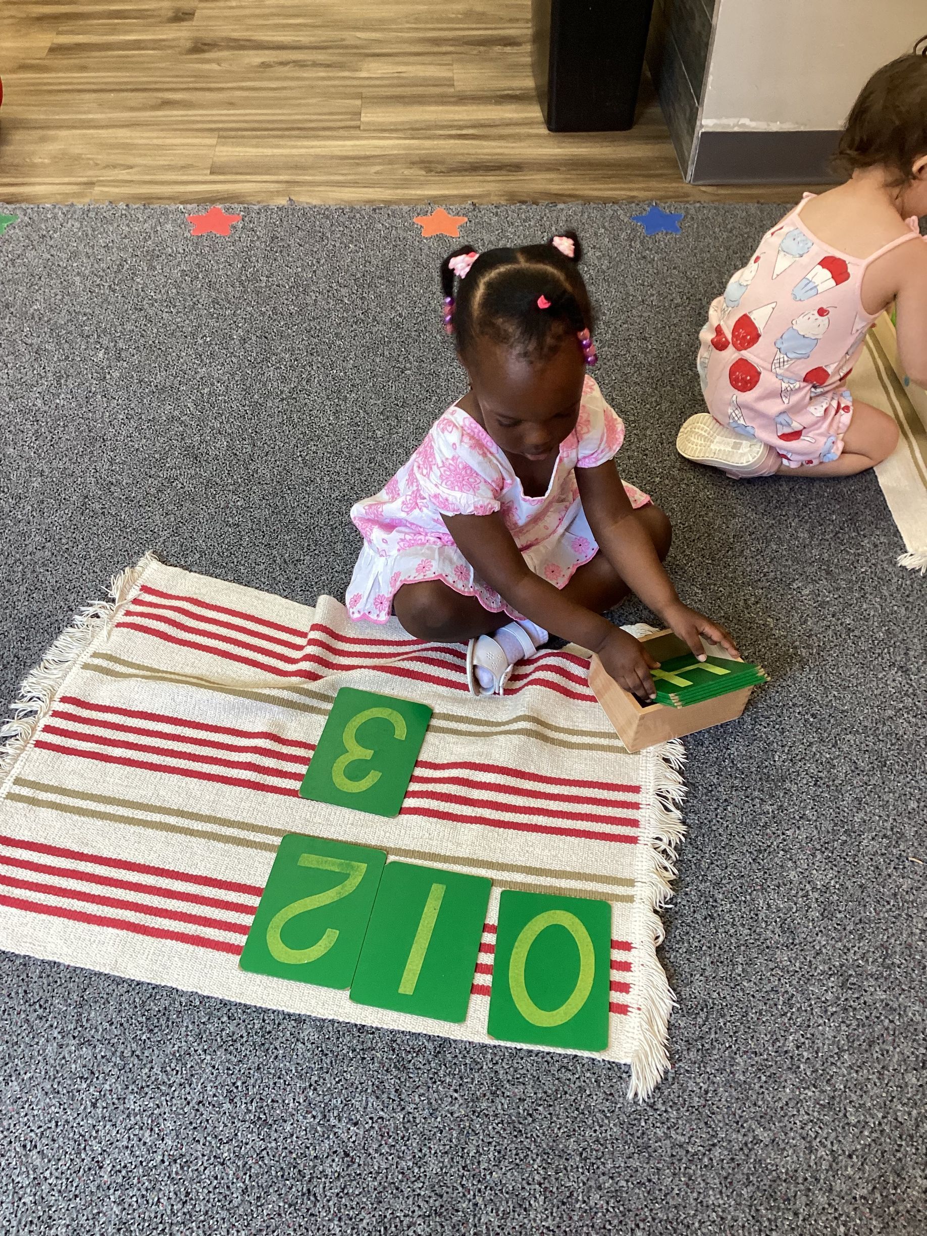 Child sitting on a rug, arranging green number cards in a classroom area.