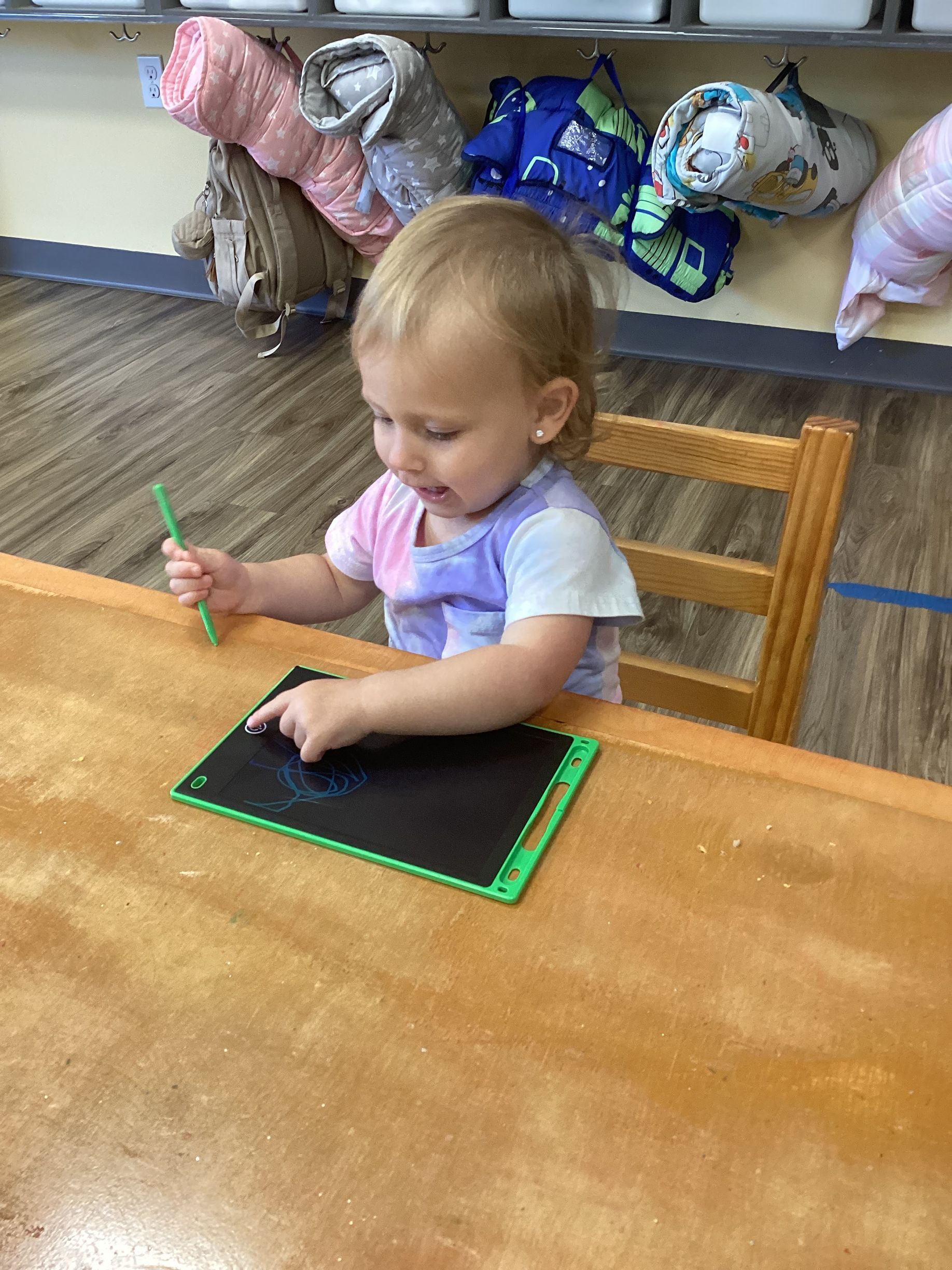 Toddler drawing on a small chalkboard at a table in a classroom, with toys on shelves behind her.