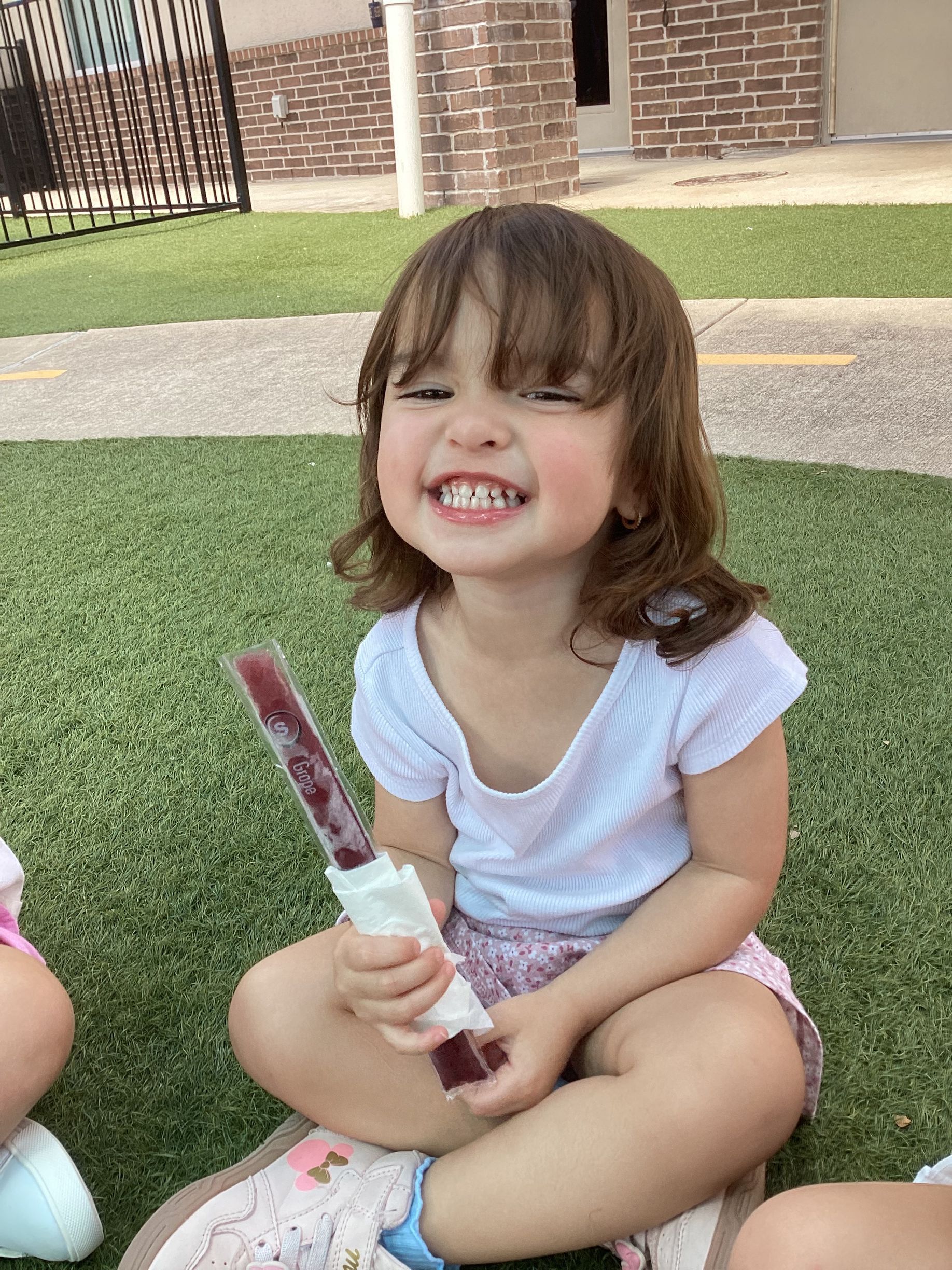Smiling child sitting on grass holding a popsicle, wearing a white shirt and pink shorts outdoors.