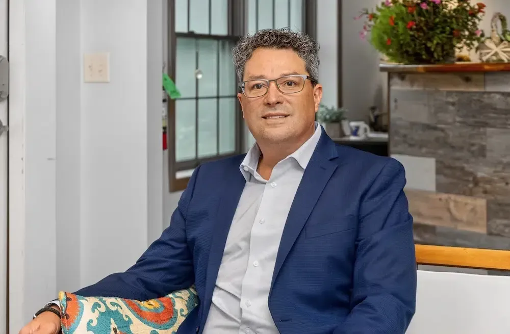 Man seated in a blue blazer in a bright living room, smiling beside a colorful cushion.