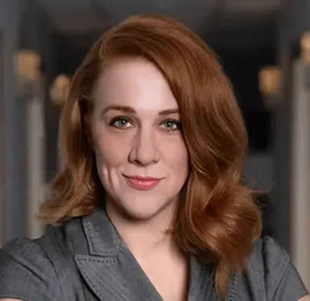 Portrait of a smiling person with auburn hair in a gray blazer against a blurred indoor background