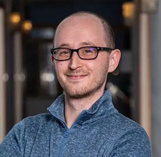 Smiling bald man with glasses in a blue sweater, standing indoors with a blurred background
