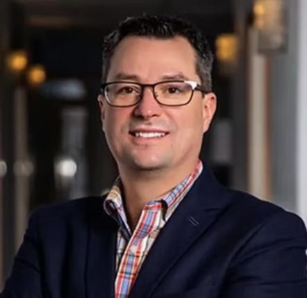 Portrait of a smiling man in glasses and a dark blazer over a plaid shirt, standing indoors against a blurred background