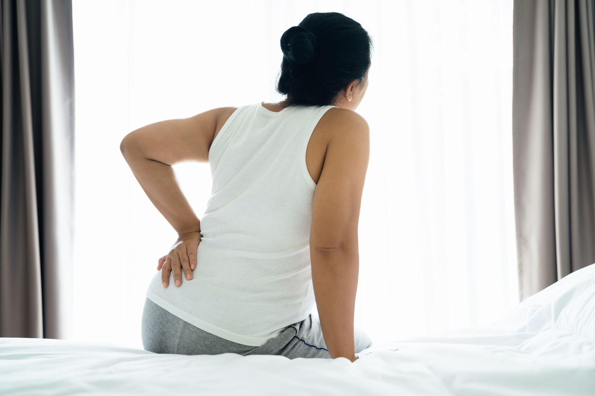 Person sitting on a bed, holding their lower back in pain near a bright window.
