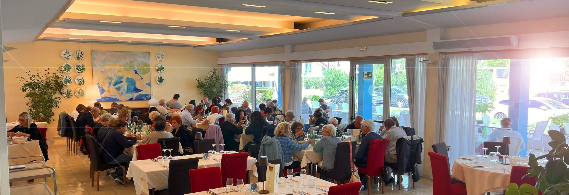 Interno del ristorante con molte persone sedute ai tavoli, grandi finestre e opere d'arte.