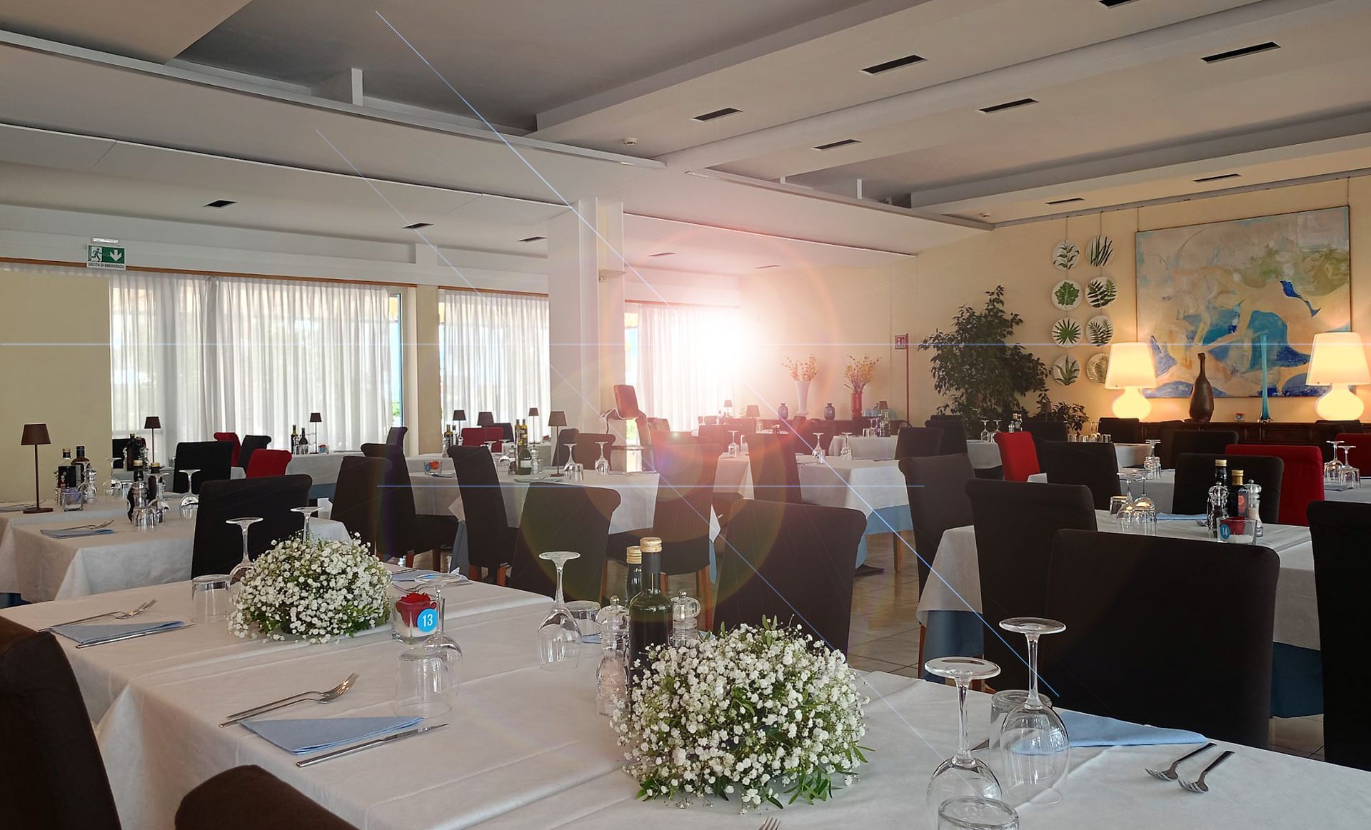 Sala da pranzo con tavoli apparecchiati per il pasto, luce solare intensa che filtra dalla finestra.