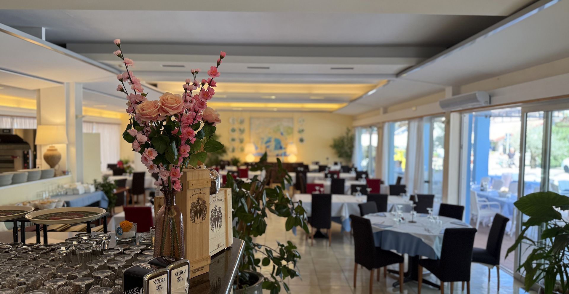 Interno del ristorante vuoto con tavoli, sedie e composizioni floreali. Le ampie finestre lasciano entrare la luce del giorno.