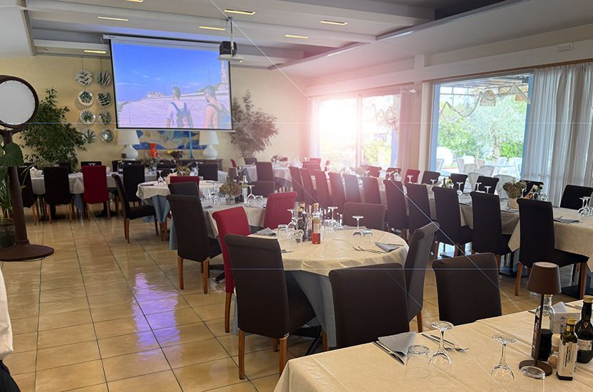 Interno di un ristorante con tavoli apparecchiati per un pasto. Lo schermo mostra un video. La luce del sole filtra da una finestra.
