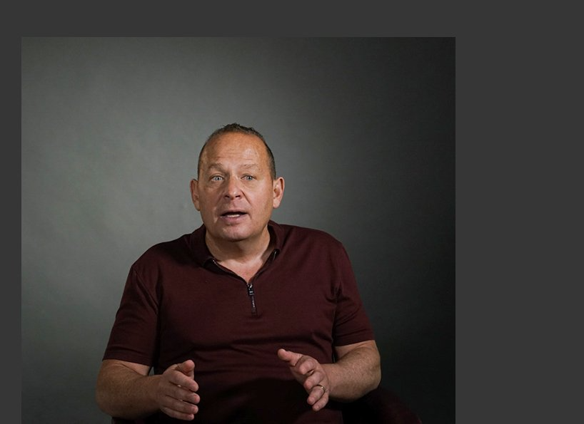 A man in a maroon shirt is sitting in front of a gray wall and talking.