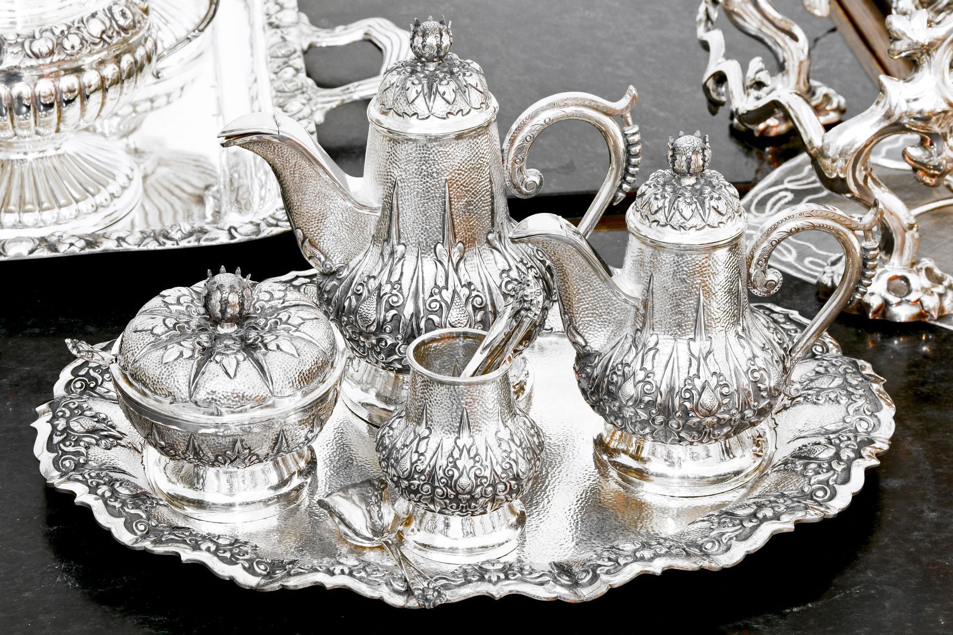 Silver tea set on ornate tray with teapot, sugar bowl, cream jug, and cups on a black table
