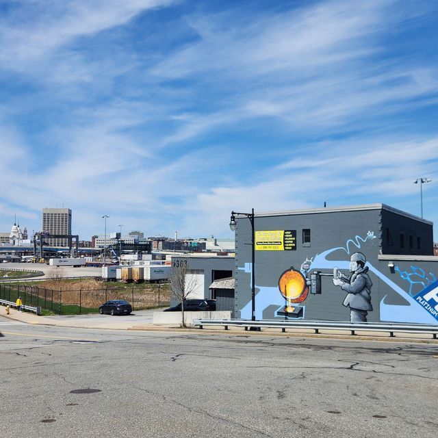 Graffiti mural on a gray building in an urban lot, with a robot painting a glowing light bulb under a blue sky