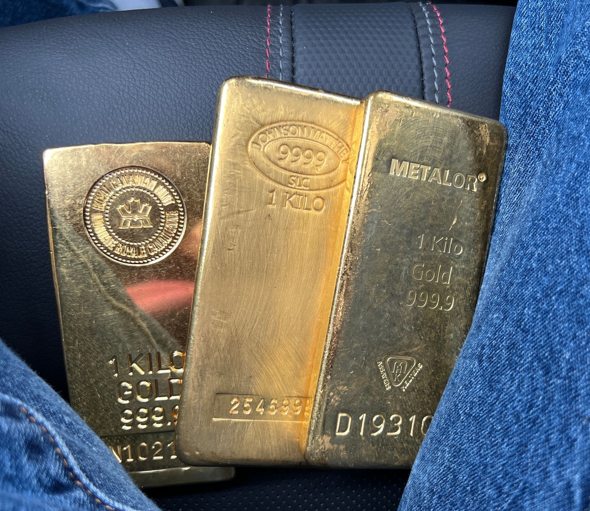 Three gold bars with stamped markings resting on a denim-clad lap.