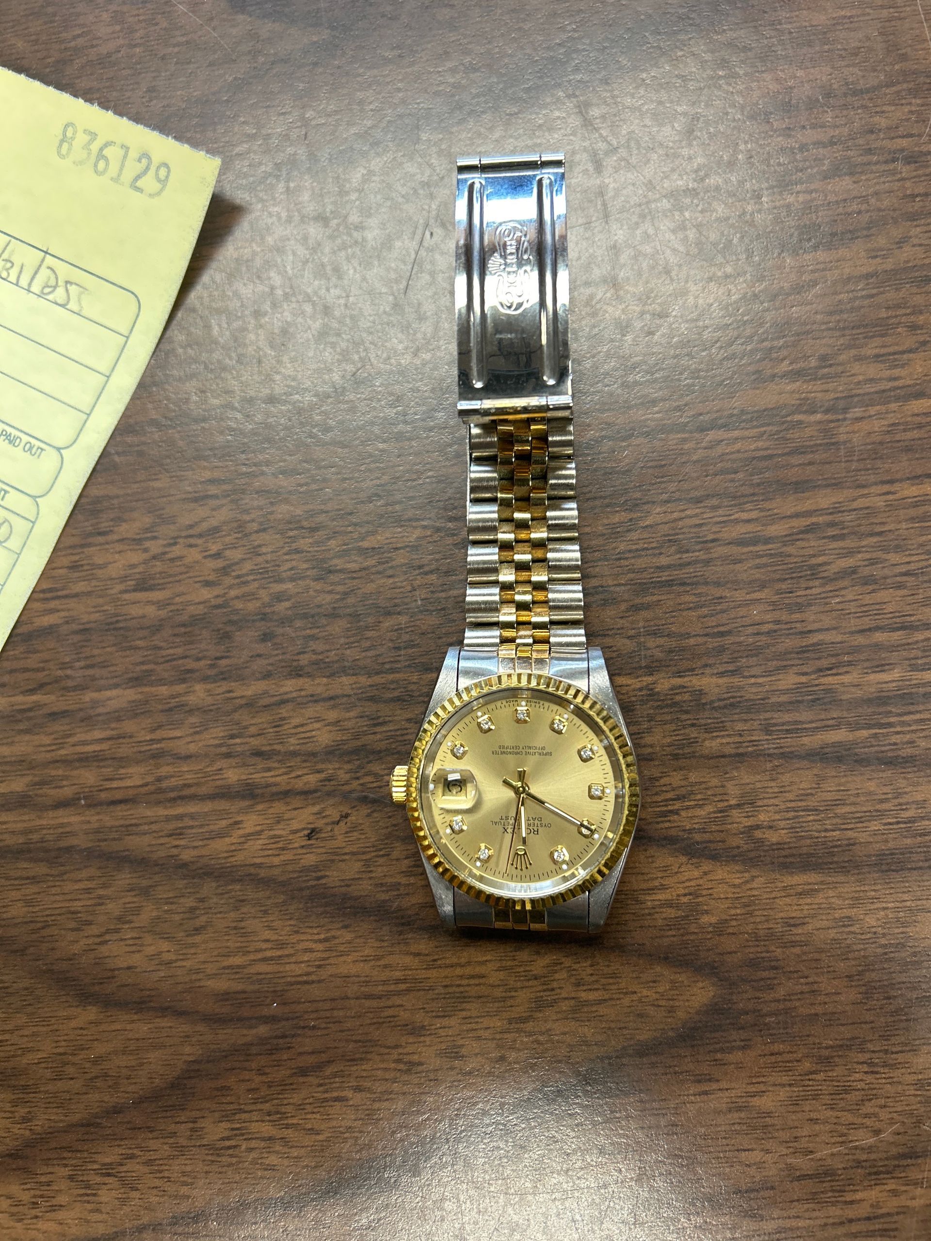 Gold-tone wristwatch with a linked band on a wooden table, next to a paper note.