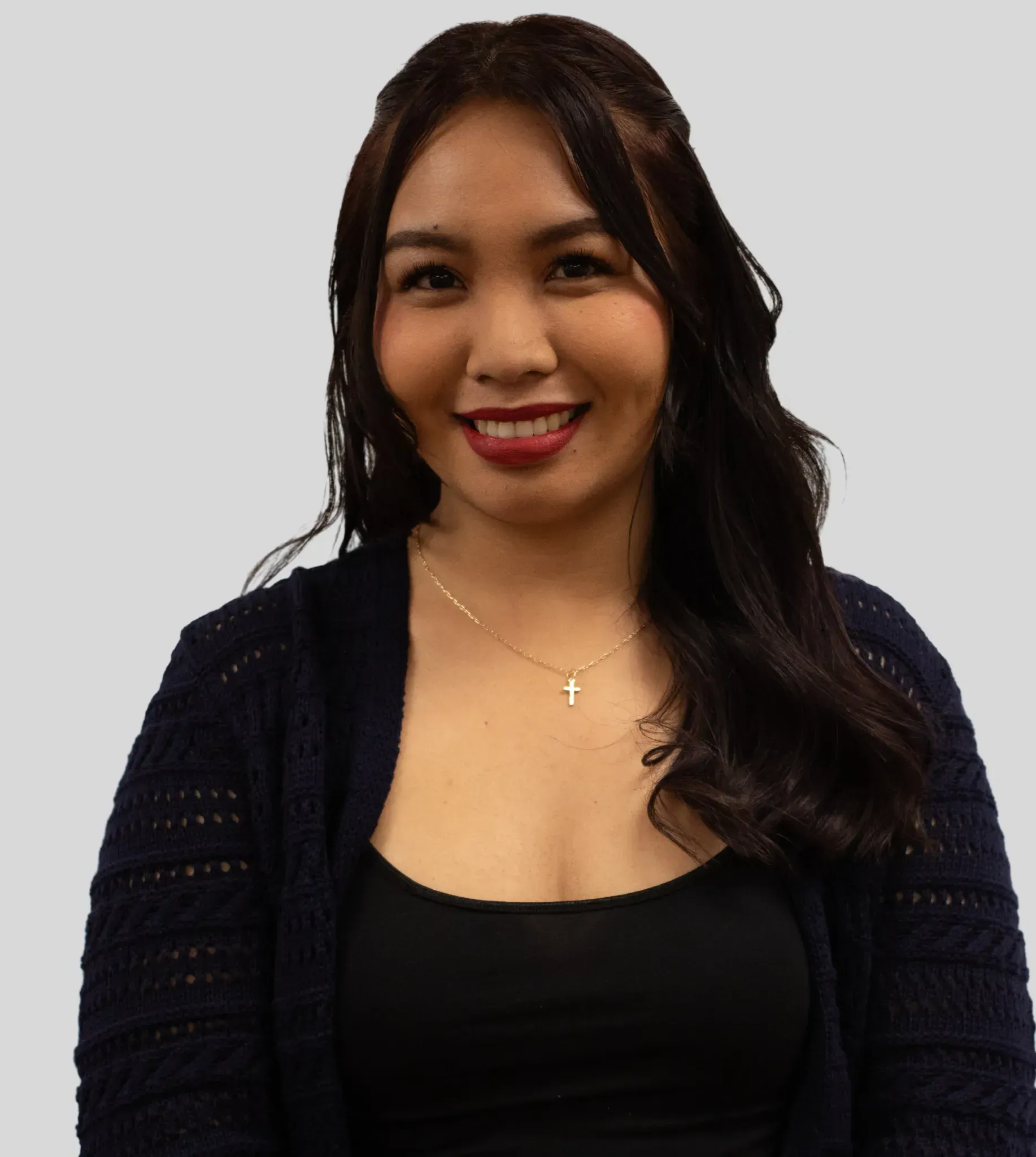 A person with long dark hair, wearing a black top and navy knit cardigan, smiles while looking at the camera.