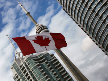 A Canadian flag waves in the foreground with the CN Tower and high-rise buildings visible in the background under a blue sky.