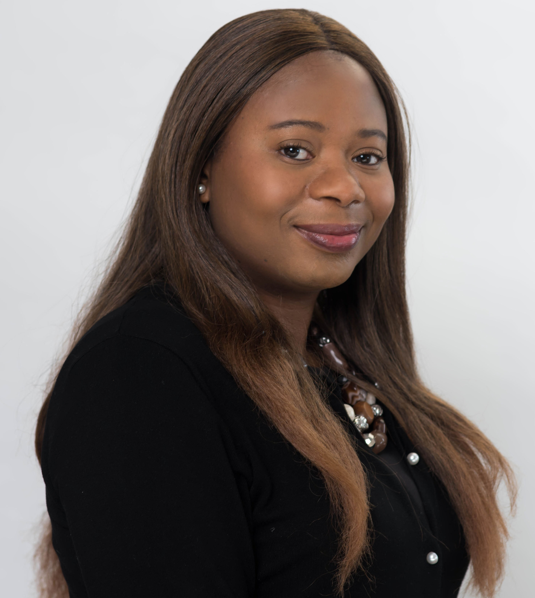 A professional headshot of a person with long brown hair, wearing a black top and beaded necklace against a white background.